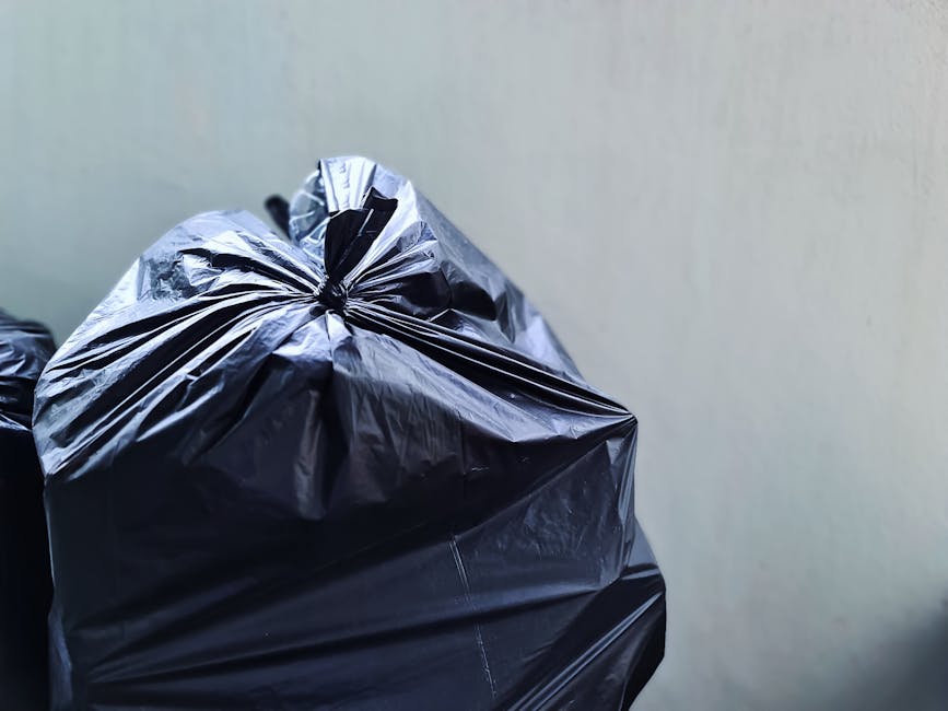 Close-up of a tightly packed black plastic rubbish bag with glossy, crinkled texture, sitting upright against a plain, light-colored wall. The bag is situated on a surface that is not fully visible bu